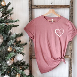 May include: A dusty rose t-shirt with a white heart design that says "Adopt" inside the heart. The heart is outlined with paw prints.