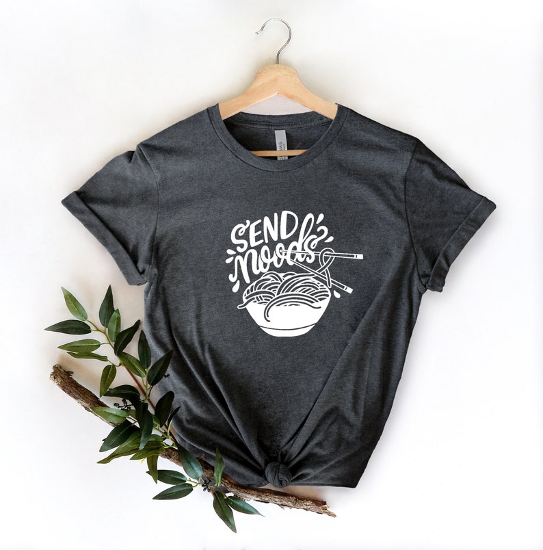 May include: A dark gray t-shirt with a white graphic of a bowl of noodles and chopsticks. The text "SEND NOODS" is written in a stylized font above the bowl.