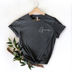 May include: Dark gray t-shirt with the word "Groom" in elegant white script. The shirt is displayed on a wooden hanger, with a knotted bottom and a sprig of greenery for decoration. A wedding-themed apparel item.