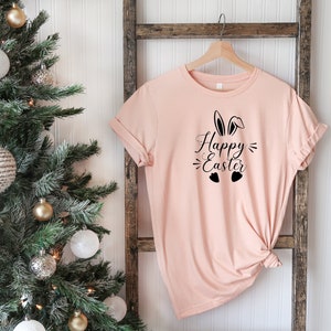 Op de afbeelding: Een lichtroze T-shirt met een zwarte afbeelding van konijnenoren en de tekst "Happy Easter" met twee hartjes eronder.