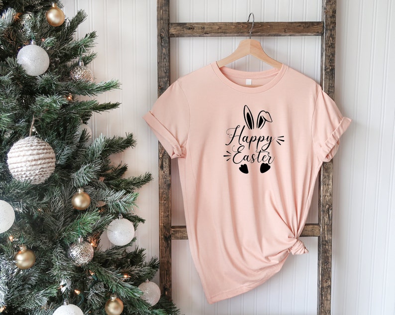 Op de afbeelding: Een lichtroze T-shirt met een zwarte afbeelding van konijnenoren en de tekst "Happy Easter" met twee hartjes eronder.