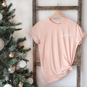 May include: A blush pink t-shirt with the word "bride" written in white script on the front.