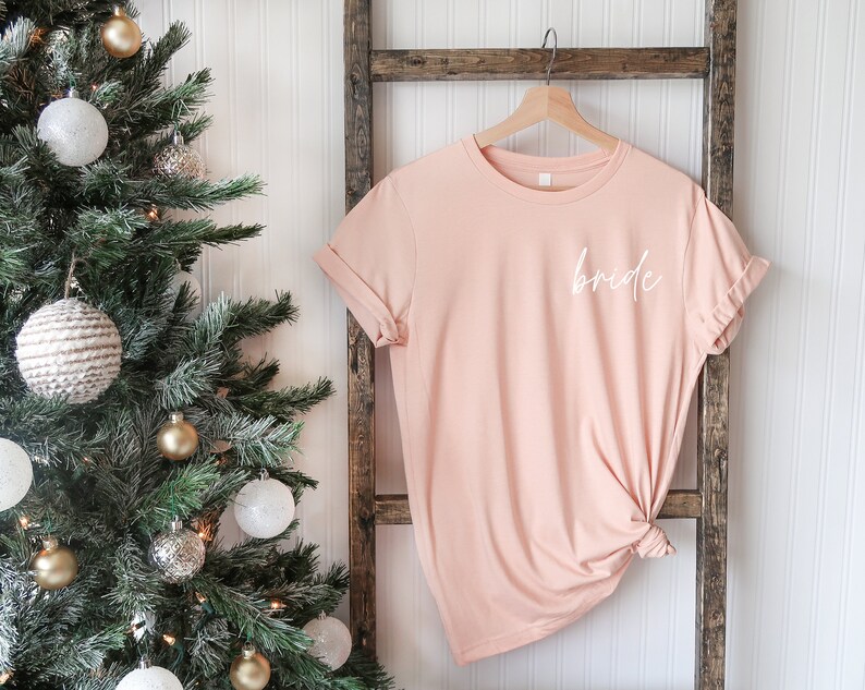May include: A blush pink t-shirt with the word "bride" written in white script on the front.