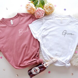 May include: Two t-shirts, one dusty rose and one white, with the words "Bride" and "Groom" in cursive script. The shirts are on a white surface with confetti and flowers. A bottle of sparkling wine is also present.