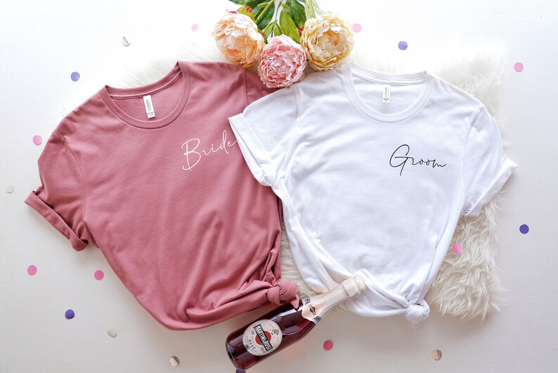 May include: Two t-shirts, one dusty rose and one white, with the words "Bride" and "Groom" in cursive script. The shirts are on a white surface with confetti and flowers. A bottle of sparkling wine is also present.