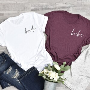 Op de afbeelding: Twee T-shirts, een wit met het woord "bride" in zwarte letters en een bordeauxrood met het woord "babe" in witte letters.