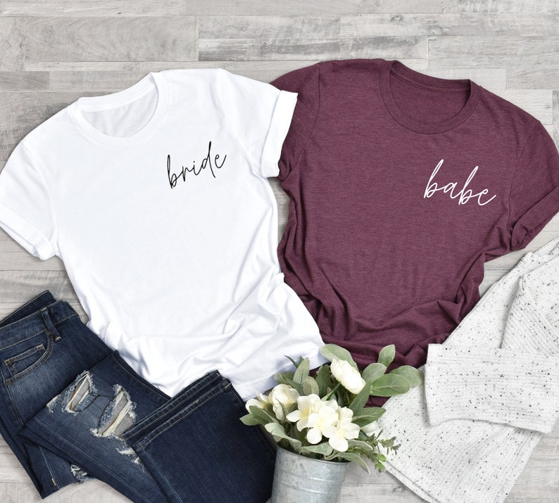 Op de afbeelding: Twee T-shirts, een wit met het woord "bride" in zwarte letters en een bordeauxrood met het woord "babe" in witte letters.