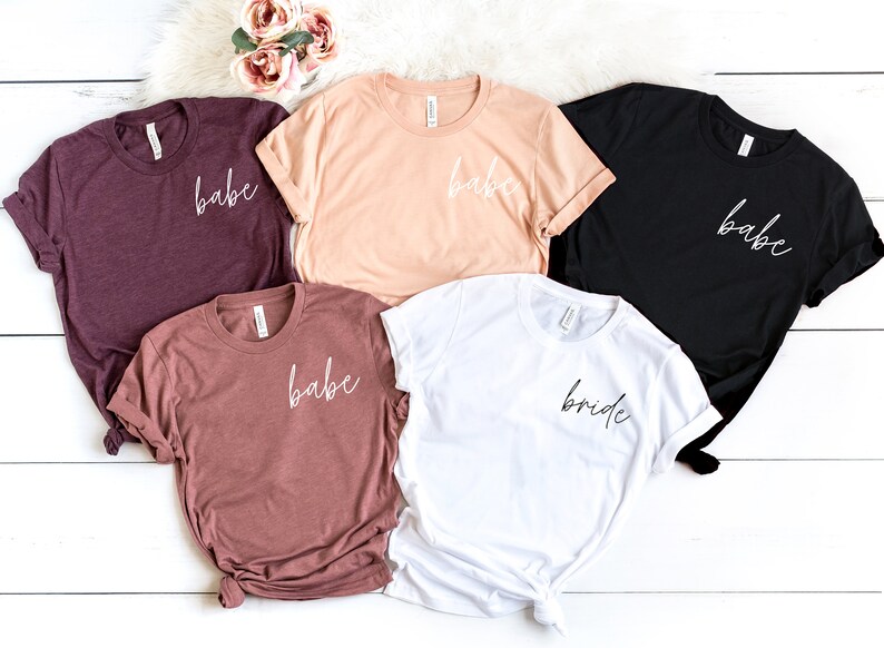 Op de afbeelding: Vijf T-shirts met de woorden "babe" en "bride" geschreven in een schrijflettertype. De shirts zijn in verschillende kleuren, waaronder bordeauxrood, perzik, wit en zwart.