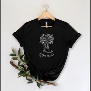 May include: Black t-shirt with a white graphic of a cowboy boot filled with sunflowers. The words "Stay Wild" are written below the boot in a script font. The shirt is hanging on a wooden hanger.