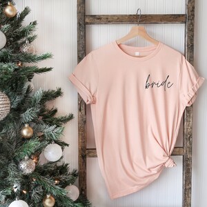 May include: A blush pink t-shirt with the word "bride" written in black script on the front.