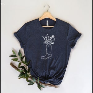 May include: A navy blue t-shirt with a white outline of a cowboy boot filled with white flowers. The tee is displayed on a wooden hanger. A sprig of greenery is placed on the lower left against a neutral background.