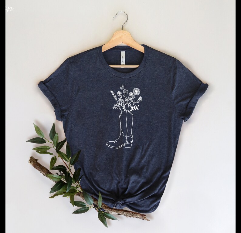 May include: A navy blue t-shirt with a white outline of a cowboy boot filled with white flowers. The tee is displayed on a wooden hanger. A sprig of greenery is placed on the lower left against a neutral background.
