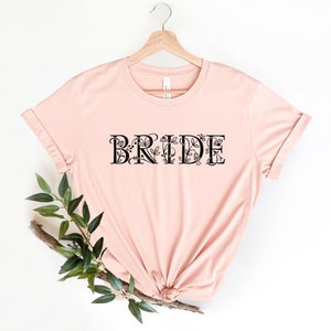 Może przedstawiać: R&oacute;żowy T-shirt z napisem "BRIDE" w czarnym kolorze z kwiatowymi akcentami. Koszulka jest zawieszona na drewnianym wieszaku.