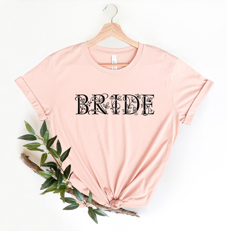 Może przedstawiać: R&oacute;żowy T-shirt z napisem "BRIDE" w czarnym kolorze z kwiatowymi akcentami. Koszulka jest zawieszona na drewnianym wieszaku.