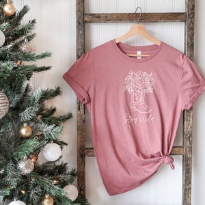 May include: A dusty rose t-shirt with a white graphic of a cowboy boot filled with sunflowers and the words "Stay Wild" in a script font. The shirt is hanging on a wooden hanger.