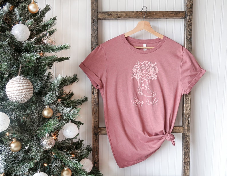 May include: A dusty rose t-shirt with a white graphic of a cowboy boot filled with sunflowers and the words "Stay Wild" in a script font. The shirt is hanging on a wooden hanger.