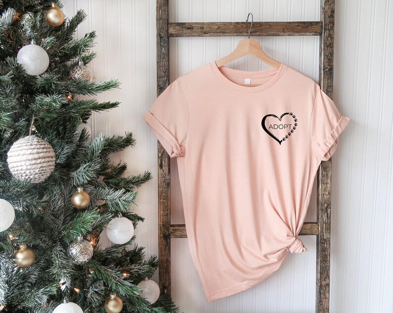 May include: A light pink t-shirt with a black heart design that says "Adopt" inside the heart. The heart is outlined with paw prints.