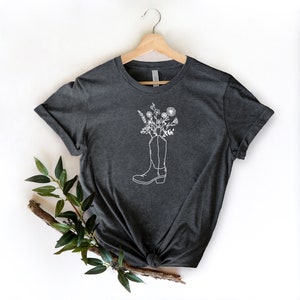 May include: Dark grey t-shirt with a white outline of a cowboy boot filled with flowers. The tee is displayed on a wooden hanger against a white backdrop. A sprig of greenery is placed on the lower left of the image.