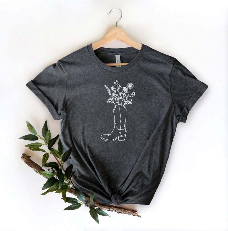 May include: Dark grey t-shirt with a white outline of a cowboy boot filled with flowers. The tee is displayed on a wooden hanger against a white backdrop. A sprig of greenery is placed on the lower left of the image.