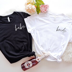May include: Two white t-shirts with black text. One shirt says "babe" and the other says "bride".