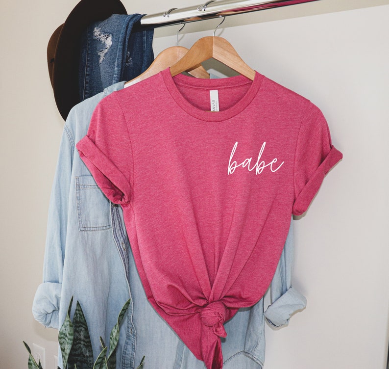 Op de afbeelding: Een roze T-shirt met het woord "babe" in wit cursief op de voorkant.