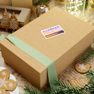 May include: A brown gift box with a green ribbon and a sticker that says "Alghero, Sardegna, Italia". The sticker has a picture of a sunset over the ocean.