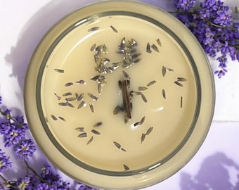 Puede incluir: Una vela en un tarro de vidrio transparente con una cera de color beige claro. La cera tiene flores y capullos de lavanda secos incrustados.