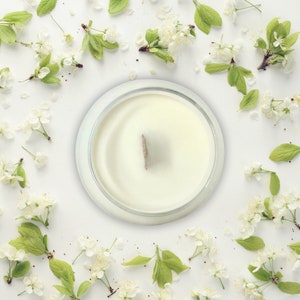 May include: A white candle in a clear glass jar with a wooden wick, surrounded by white blossoms and green leaves on a white background.