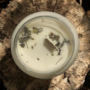 May include: A white candle with dried herbs and a small piece of wood inside a clear glass jar. The candle is sitting on a wooden surface.