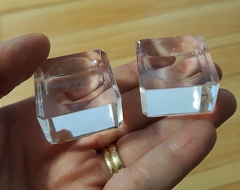 Puede incluir: Dos candelabros cuadrados de vidrio transparente con base rectangular se sostienen en una mano. Los candelabros tienen un diseño facetado y son transparentes, reflejando la luz. El fondo es una superficie de madera.