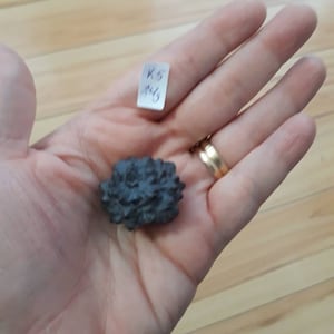 May include: A small, dark grey, rock-like cluster of crystals. The cluster is about the size of a 20 pence coin and is being held in a hand. A small white label with the text "K5 1/4" is visible in the background.