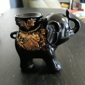 May include: Black elephant figurine with a gold decorative design on its back. The elephant is holding a small, round, black bowl on its back.