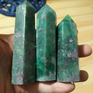 May include: Three green and pink crystal points, also known as green phantom quartz, are held in a hand. The points are different sizes and have a natural, rough texture.