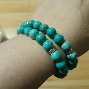 May include: Two turquoise beaded bracelets with silver accents. The bracelets are stacked on a person's wrist.