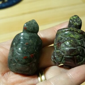 Puede incluir: Dos figuras de tortugas de piedra tallada. Las tortugas son de color verde oscuro con detalles rojos y verdes. Una tortuga tiene un acabado liso, mientras que la otra tiene un caparazón texturizado. Las figuras son pequeñas y parecen talladas a mano.