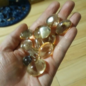 May include: A hand holding a collection of small, clear, oval-shaped glass stones with a golden hue.