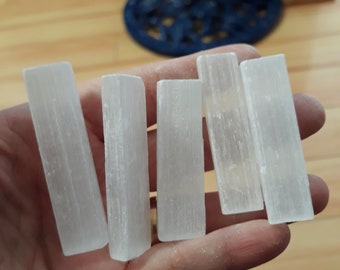 Op de afbeelding: Vijf rechthoekige, doorschijnende witte selenietkristalstaven worden in een hand gehouden. De staven hebben een glad, licht getextureerd uiterlijk. De achtergrond is wazig en toont een houten vloer en speelgoed.