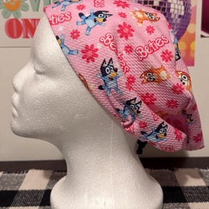 May include: Pink surgical scrub hat with a repeating pattern of cartoon dogs and flowers. The text "Betties" is printed on the hat.