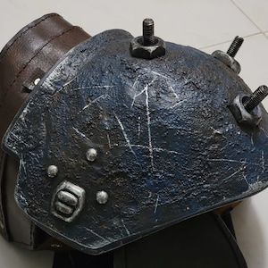 May include: A blue and brown knee armor with metal accents and bolts. The armor is made of a textured material and has a worn look.