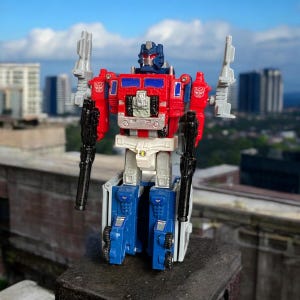 Könnte beinhalten: Ein rotes, weißes und blaues Roboter-Spielzeug mit einem LKW-Kabinen-Torso und Waffen. Der Roboter steht auf einer grauen Oberfläche mit einer Skyline im Hintergrund.