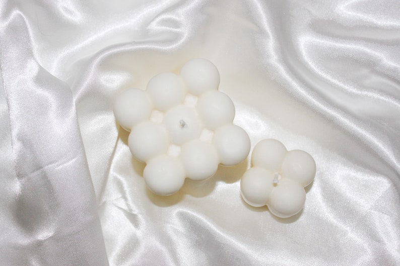 May include: Two white bubble candles, one large and one small, on a white satin background.