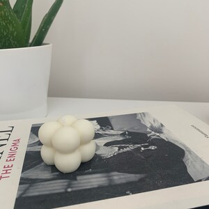 May include: A white, geometric candle with a bubble-like design. The candle is sitting on a book with a black and white photo of a person.