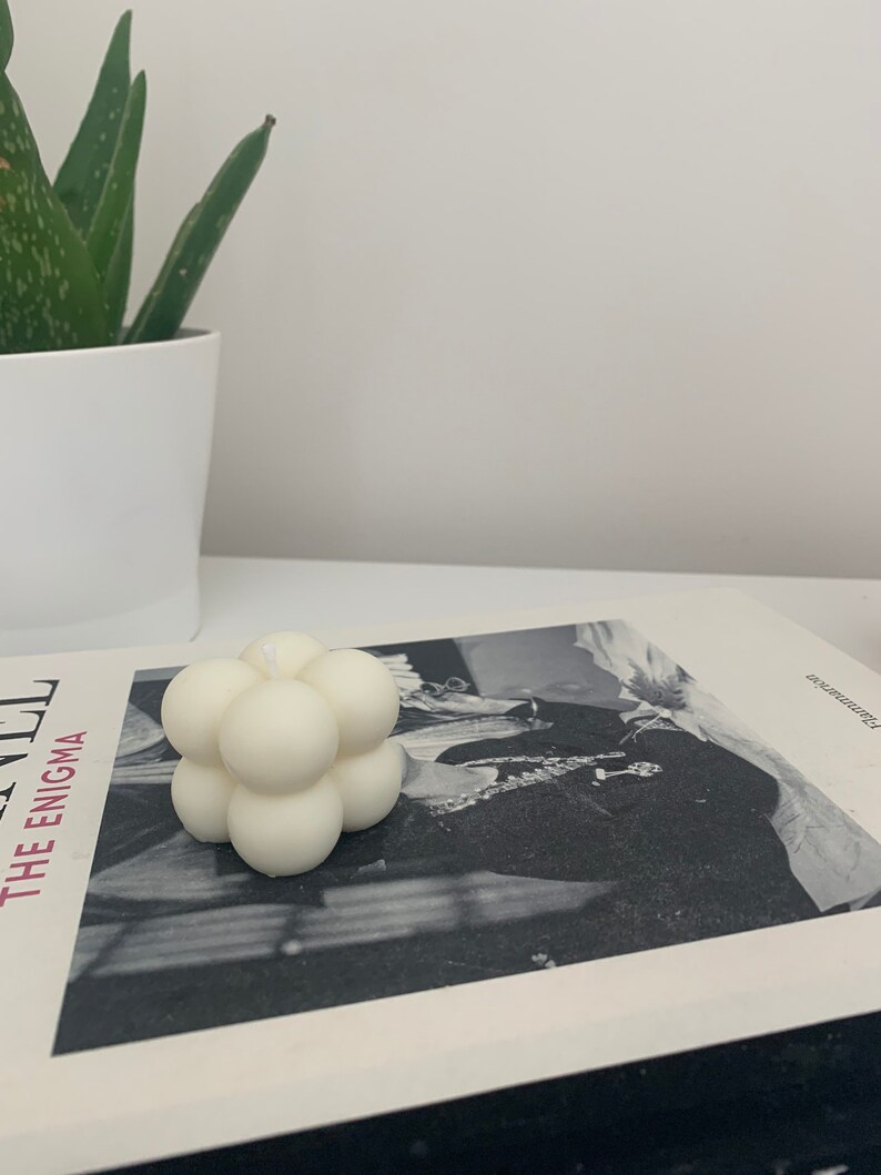 May include: A white, geometric candle with a bubble-like design. The candle is sitting on a book with a black and white photo of a person.