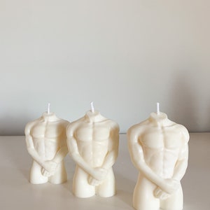 May include: Three white sculpted candles shaped like a human torso. The candles are standing on a white surface.