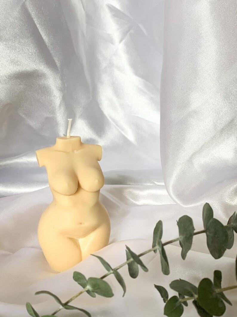 May include: A nude-colored candle shaped like a human torso. The candle is sitting on a white satin fabric with green eucalyptus leaves.