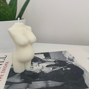 May include: A white candle shaped like a human torso. The candle is sitting on a book with a black and white photo of a woman on the cover.