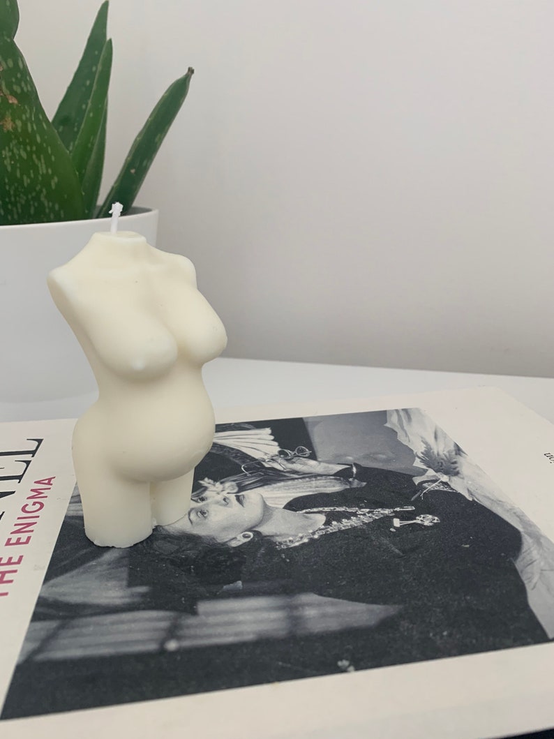 May include: A white candle shaped like a human torso. The candle is sitting on a book with a black and white photo of a woman on the cover.