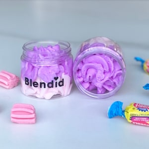 May include: Two small glass jars filled with purple and white whipped body butter. The jars are labelled "Blendid" with a heart symbol.  There are also several pink and yellow wrapped sweets on a white surface.
