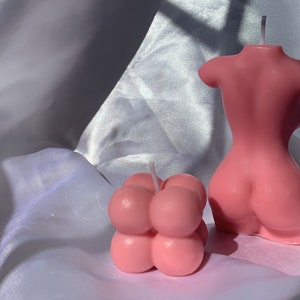 May include: Two pink candles, one shaped like a human torso and the other shaped like a cluster of bubbles.