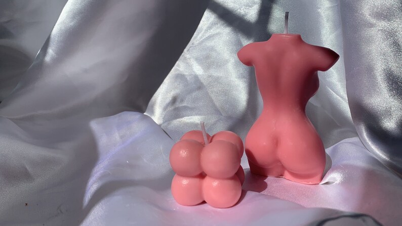 May include: Two pink candles, one shaped like a human torso and the other shaped like a cluster of bubbles.